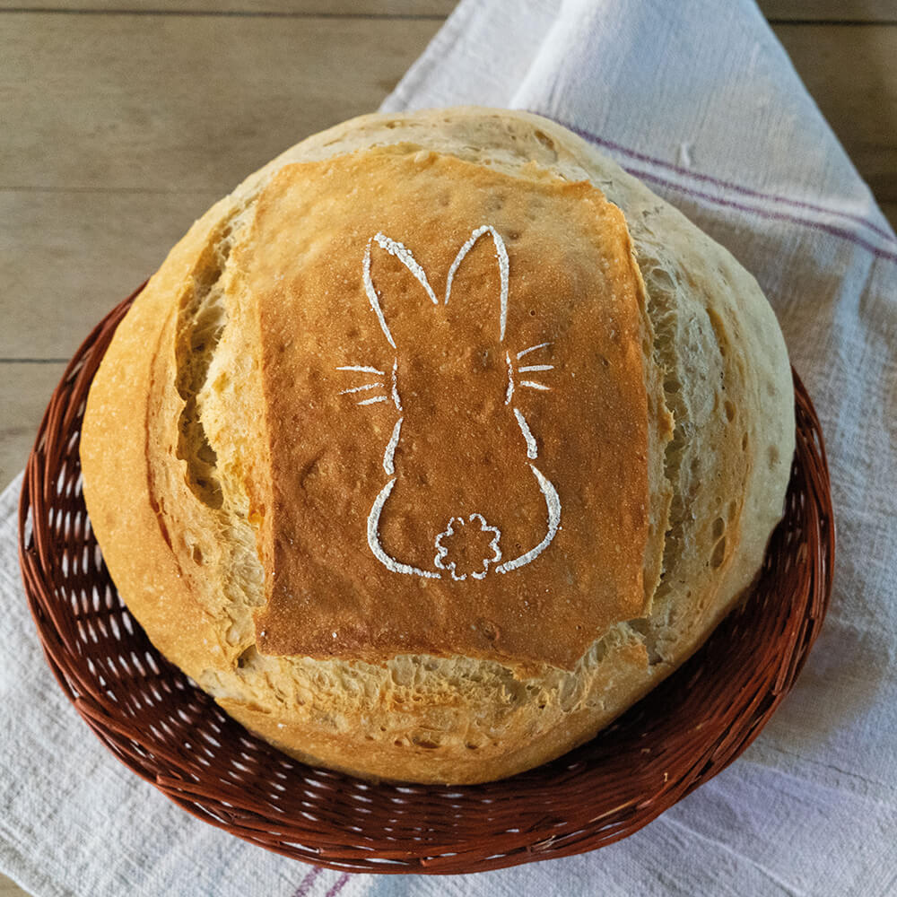 Ein Brotlaib liegt in einem geflochtenen Korb, der auf einem Tuch steht. Auf dem Brot ist ein Bild aus Mehl zu sehen in Form eines sitzenden Hasen.