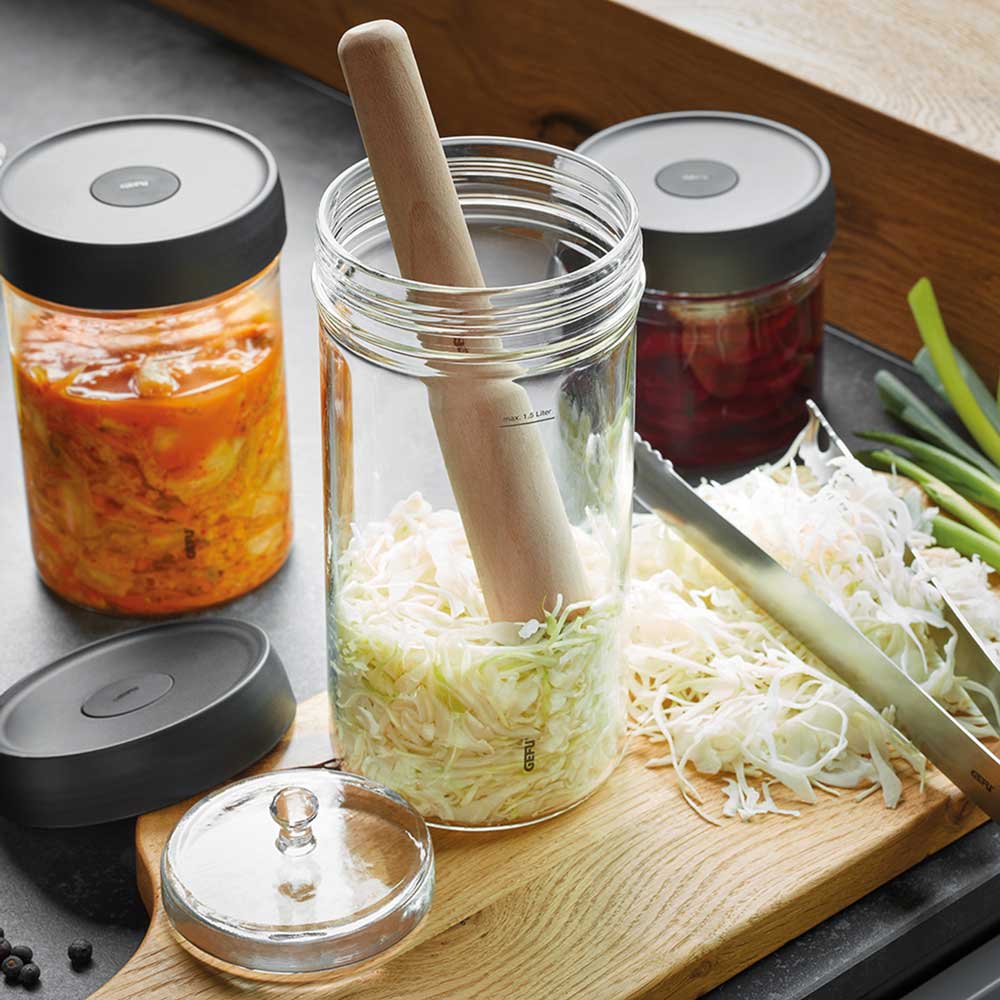 Fermentierset 4-teilig Fermentierset mit einem Holzstößel im Glas. Im Glas befindet sich eingemachter Lauch. Im Hintergrund befinden sich weitere Fermentiergläser mit fermentiertem Gemüse in einer Küche.