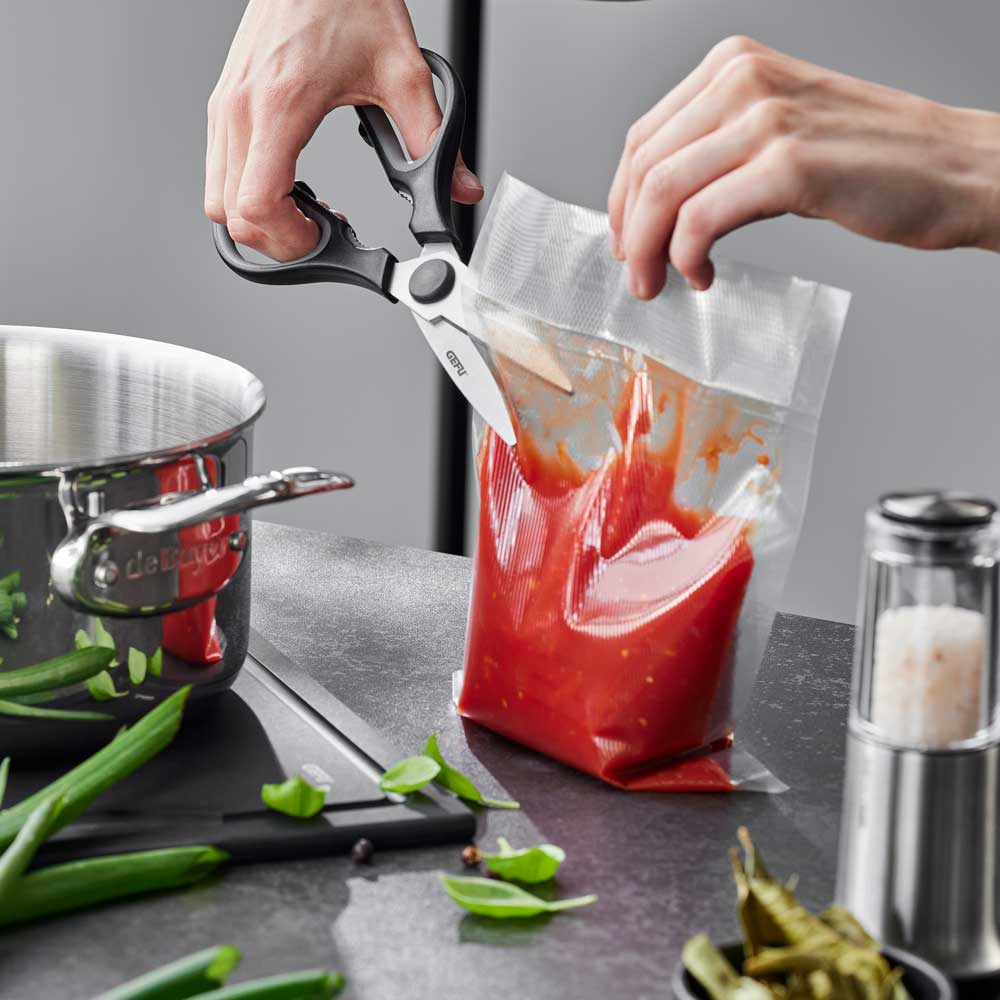 Eine Person schneidet mit der Schere einen vakumierten Beutel auf einer Küchenarbeitsplatte auf. Daneben stehen verschiedene weitere Küchenuntensilien.