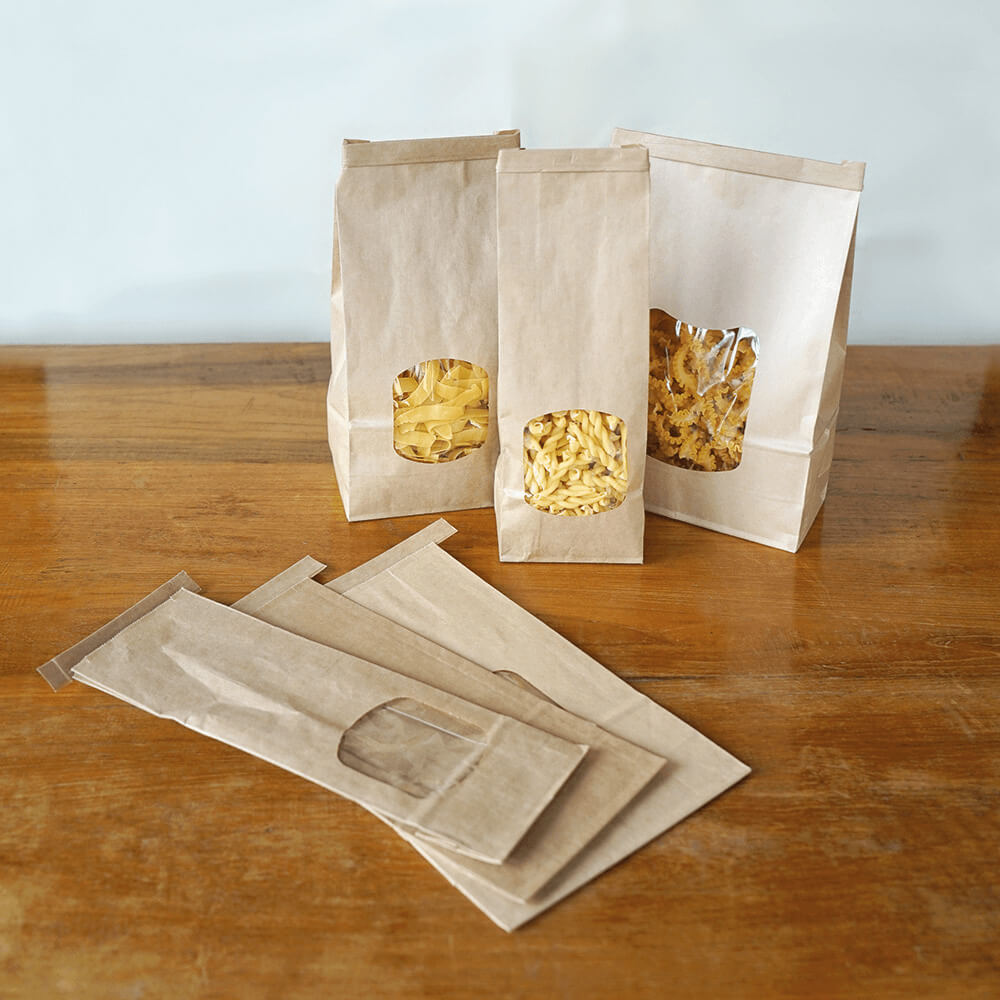 3 gefüllte Nudelverpackungen mit Fenster auf einem Holztisch. Daneben liegen 3 leere, flache Verpackungen