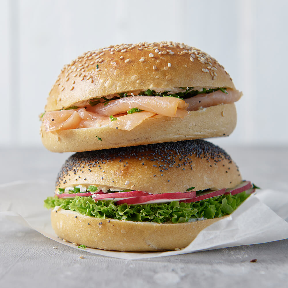 zwei Belegte Bagels, der mit Sesam ist belegt mit Lachs und Schnittlauch und der mit Mohn ist belegt mit Salat und Radieschen