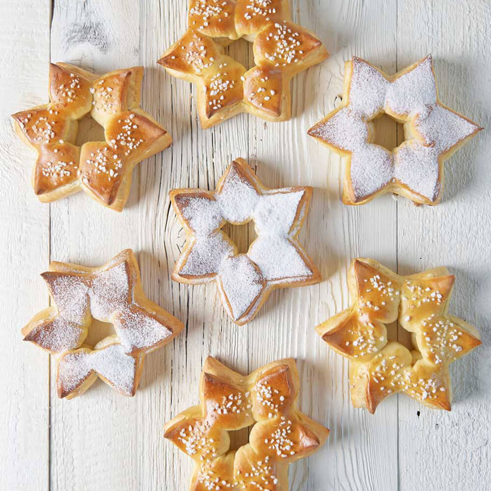 Sternbrotdrücker 7 Sternförmige Brote die alle ein Loch in der Mitte haben. Vier Sternförmige Brote sind mit Hagelzucker bestreut und drei Brote mit Puderzucker. Diese Brote siegen alle auf einer hellgrauen Oberfläche
