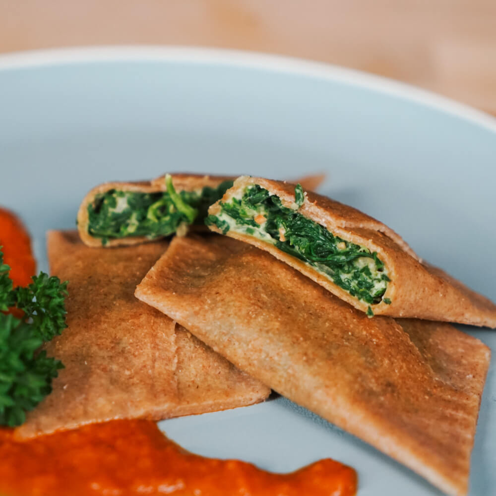 Rezept für Vollkornmaultaschen Leckere Vollkornmaultaschen mit einer Spinat-Ricotta-Füllung und einer Tomatensoße, angerichtet auf einem hellblauen Teller