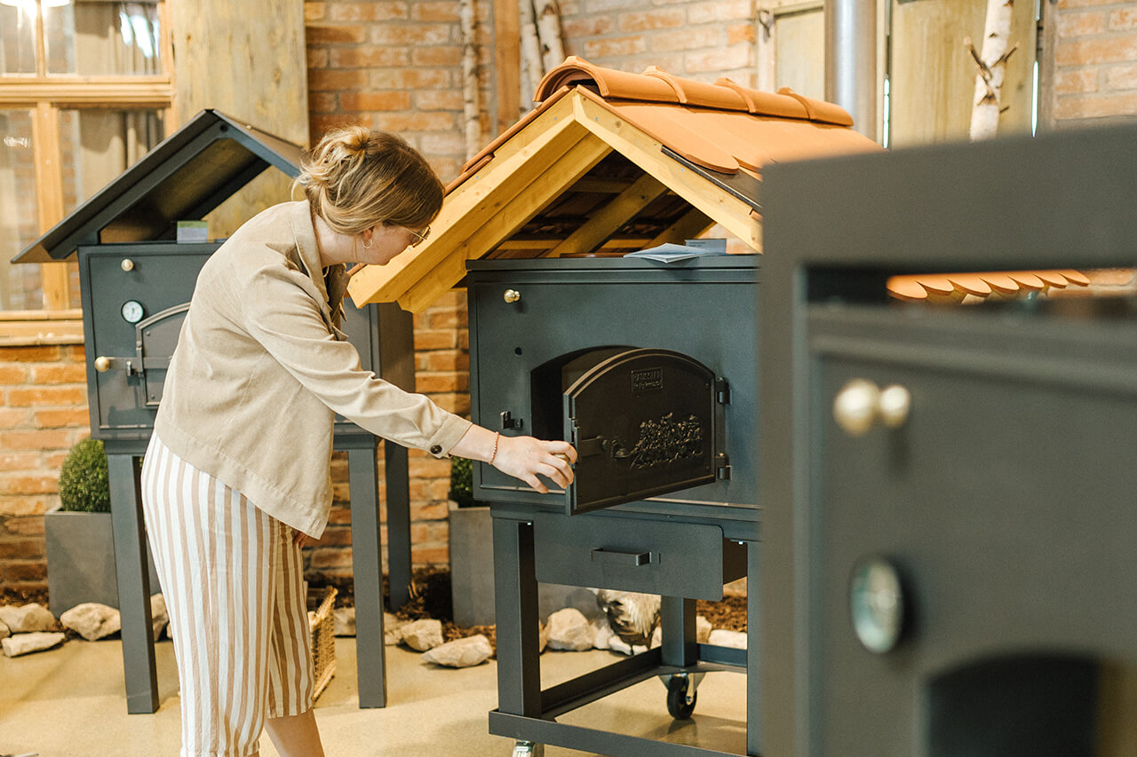 Besucherin_Holzbackofen Besucherin im Backdorf schaut neugierig in einen traditionellen Holzbackofen, umgeben von rustikaler Backstuben-Atmosphäre.