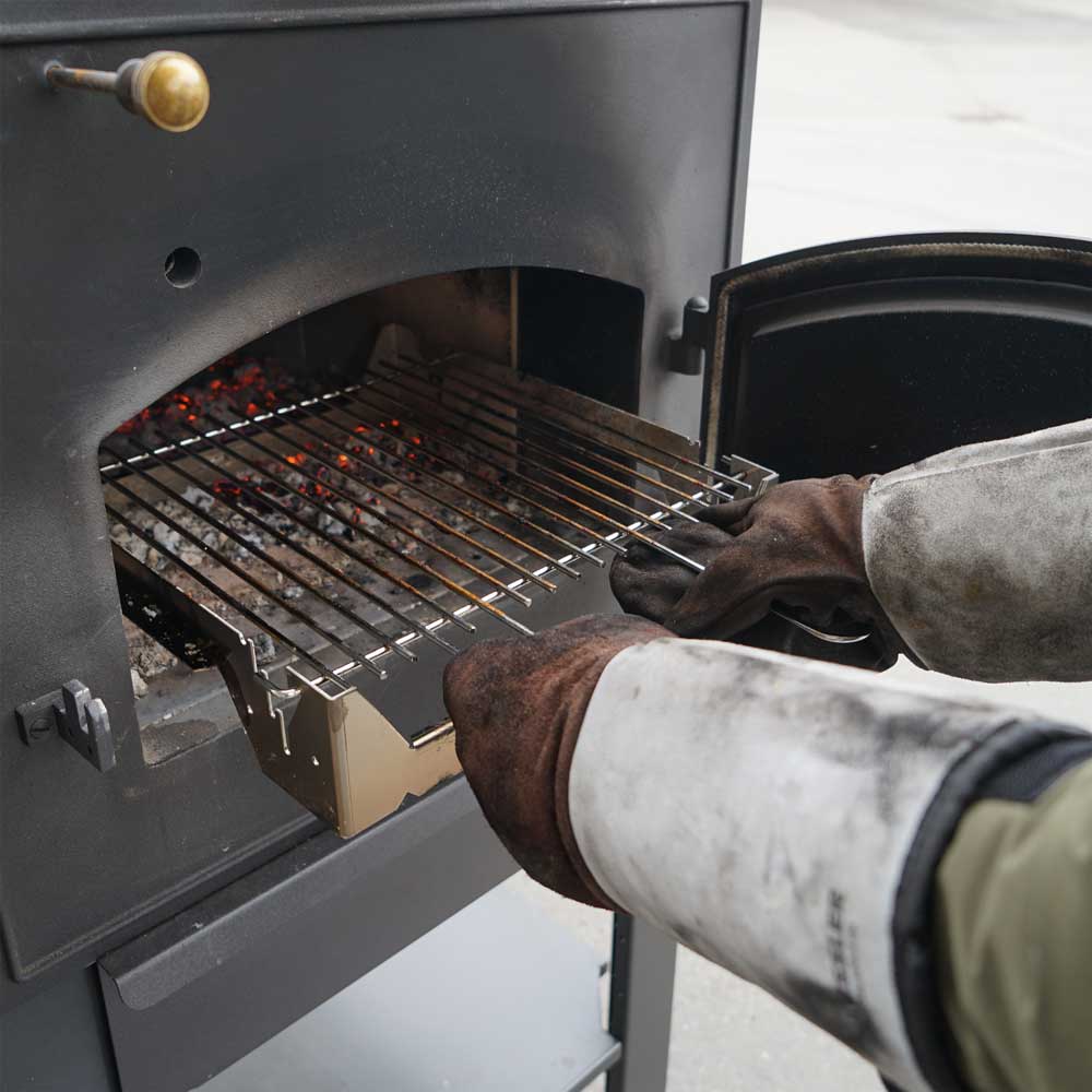 Grillrost wird in Holzbackofen geschoben. Es werden dafür Grillhandschuhe getragen.