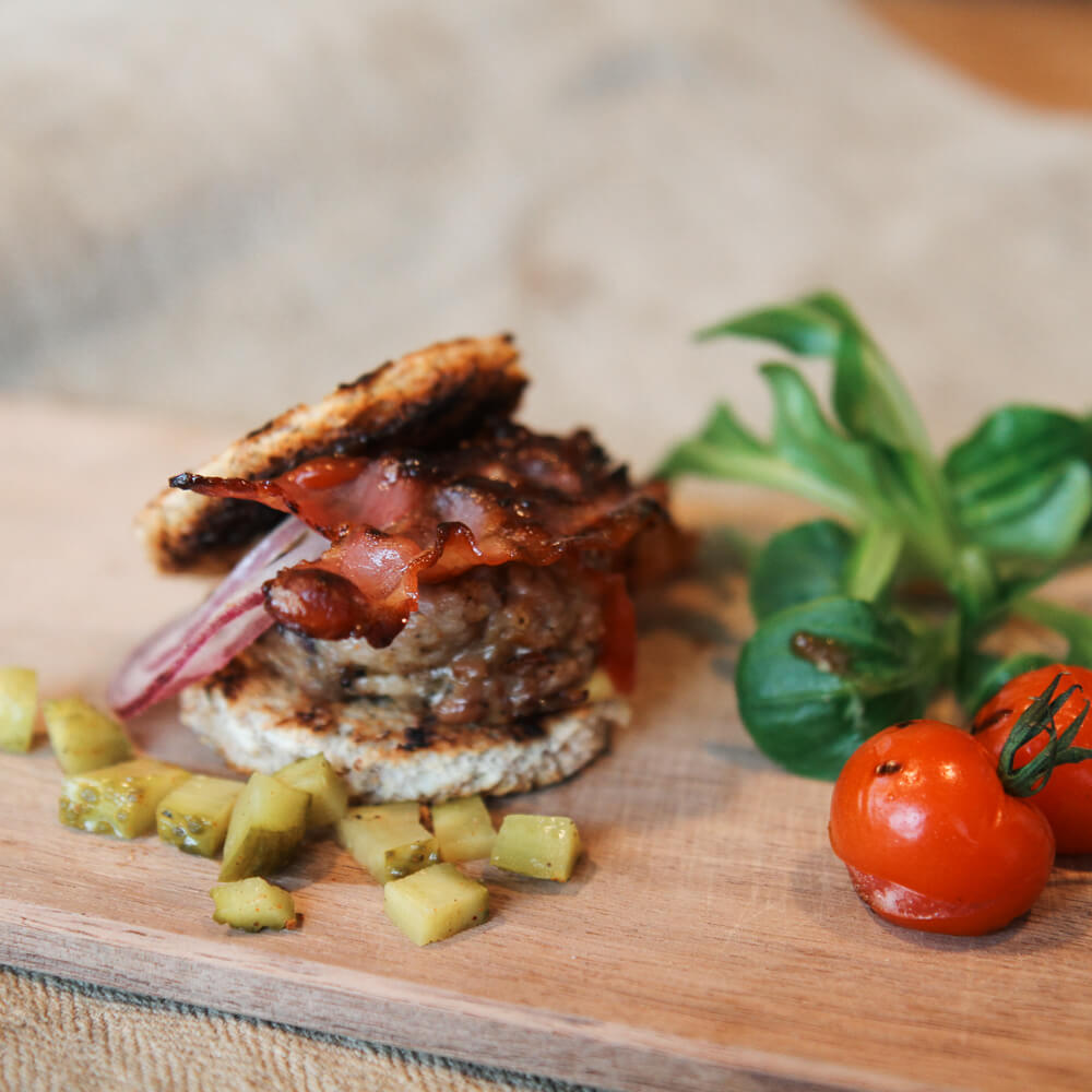 Rezept für Miniburger selbstgemachte Miniburger mit Speckscheiben und Essiggurken belegt auf einem Holzbrett serviert mit zwei Coctailtomaten und frischen Kräutern