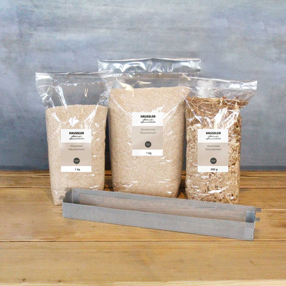 Holztisch mit 3 verschiedenen Räuchermehlen in durchsichtigen Beuteln. Davor liegt ein Smoker-Einsatz. Hintergrund grau