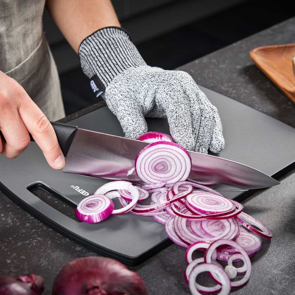 Eine Person trägt den Schnittschutzhandschuh und schneidet eine Zwiebel auf einem schwarzen Brett auf der Küchenarbeitsplatte.