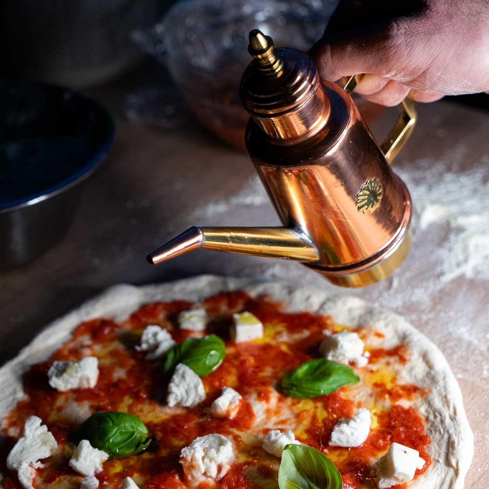 Praktische Ölkanne zum Dosieren rose- und folgdarbige Ölkanne. Gießkannenartig mit goldenem Henkel und dünnem Hals. Darauf befindet sich ein strenartiges Motiv. Auf dem Bild wird die Ölkanne benutzt um eine Pizza zu beölen. Im Hinergrund befinden sich Backutensilien