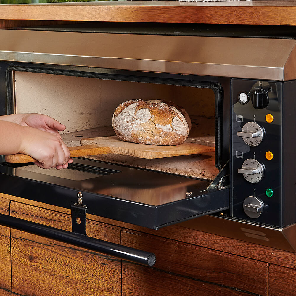 Zwei Hände entnehmen m it einem Holzschieber frisch gebackenes Brot aus einem Elektro-Steinbackofen.