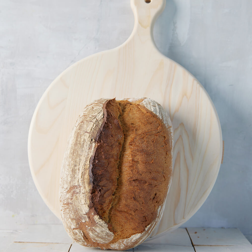 Rezept für Adelindes Schwäbisches Dinkelbrot längliches Schwäbisches Dinkelbrot aufgestellt vor einem runden Backbrett mit Griff, im Hintergrund ist eine hellgraue Wand